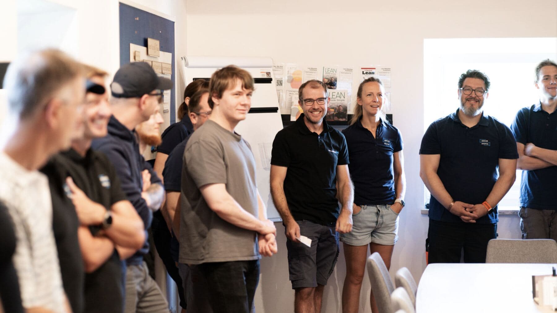 Das Tradeo-Team steht lachend im Kreis beim täglichen Morgenmeeting vor der Lean-Pinnwand